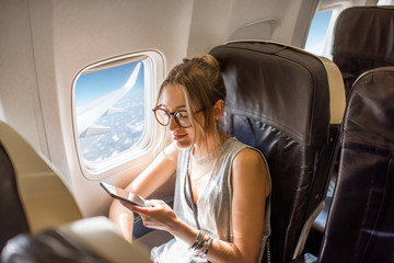 Fototapeta premium Młoda kobieta siedzi z telefonu na siedzeniu samolotu w pobliżu okna podczas lotu w samolocie