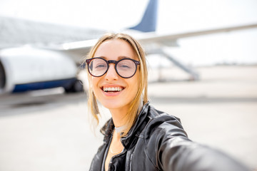 Young happy woman making selfie photo in front of the airplane on the airport runway before the...