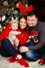 Parents play with little daughter in deer hat holding her on their arms and sitting before a Christmas tree