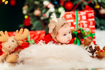 Little baby in a hat with deer lies among red present boxes before a Christmas tree