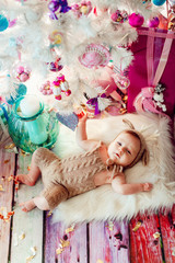 Baby in deer suit lies on soft white pillow under white Christmas tree with pink toys