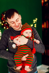 Dad holds kid in stripped suit standing before a Christmas tree