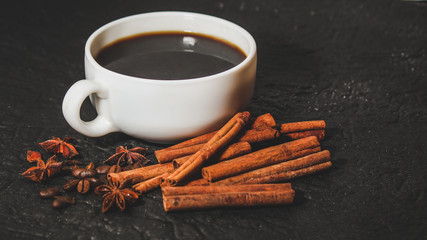 cup of coffee next to the grains of fried coffee, anise star and cinnamon sticks on black background