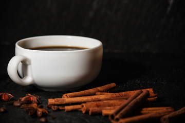 cup of coffee next to the grains of fried coffee, anise star and cinnamon sticks on black background