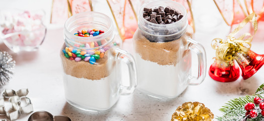 Christmas homemade gift concept, ingredients for preparation chocolate and rainbow cookies in jar, marshmallow in dish, metallic cutting forms for cookies and decorations on kitchen table