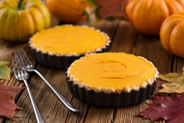 Couple of simple open pumpkin pies in baking dishes served with forks and pumpkins on rustic wooden background