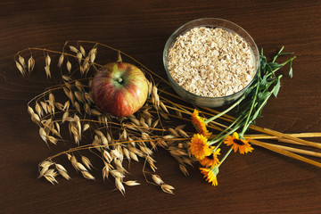 Oat ears stems and oat flakes in a white pot, marigold flowers and apple on dark brown wood table. Top view. oat flakes small size grind. Dietary breakfast