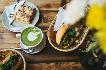 Healthy breakfast in the restaurant: cup of green tea with milk, dessert and two sandwiches with vegetables and herbs on wooden table.