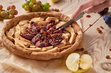 Man slices with silver vintage knife a homemade apple plums pie decorated with fresh apples, plums, grapes, brown raisins and sesame on light wooden background. Side view