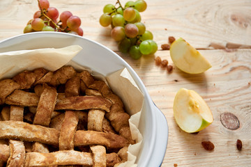 A piece of homemade american apple pie in baked form on brown wooden table with raisins and fresh fruits - apples, grapes on brown wooden background. Top view