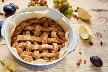 Just cooked homemade american apple pie in baked form on brown wooden table with raisins and fresh fruits - apples, grapes, plums on brown wooden background. Top view