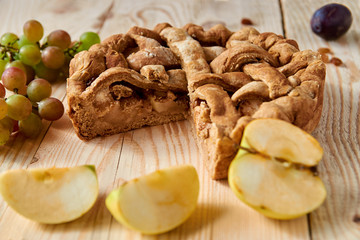 Sliced homemade american apple pie on brown wooden table with raisins and fresh fruits - apples, plums and grapes on blurred background. Side view
