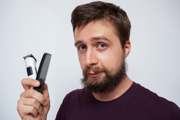 Young bearded man holding up a beard trimmer and comb while smiling
