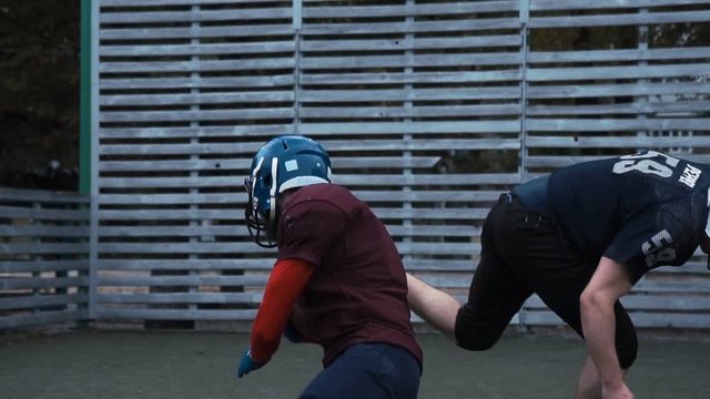 Slow motion of two helmeted football players in two different uniforms pushing past each other with fence in background
