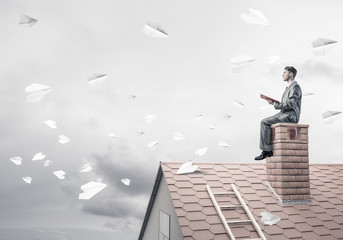 Man on brick roof reading book and paper planes flying in air