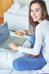 a young smiling woman in jeans and a white sweater sitting on the bed with a laptop computer and holds in her hand a credit card.