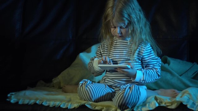 A little girl in striped pajamas sits on the sofa at night in her room and uses a smartphone, promising her parents or a nanny that she will go to bed on time.