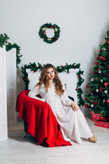 Obraz na płótnie Canvas Beautiful girl in a white dress is sitting near a Christmas tree.