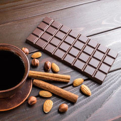Hot black coffee in brown clay cup, dark chocolate bar, almond, hazelnut and cinnamon stick on wooden background