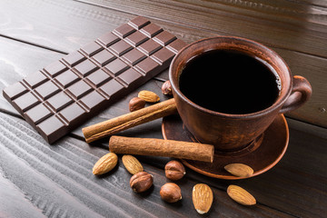 Hot black coffee in brown clay cup, dark chocolate bar, almond, hazelnut and cinnamon stick on wooden background