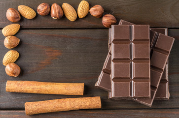 Bitter, dark chocolate bar, almond, hazelnut and cinnamon stick on wooden background with space for text, top view