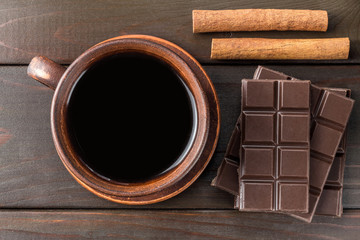 Hot black coffee in brown clay cup, dark chocolate bar and cinnamon stick on wooden background, top view