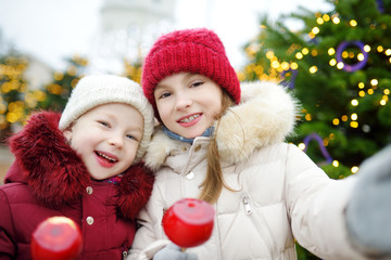 Two adorable little sisters eating red apples covered with sugar icing on traditional Christmas market