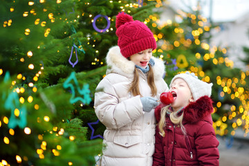 Two adorable little sisters eating red apples covered with sugar icing on traditional Christmas market