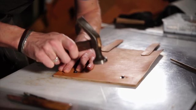 Close up shot of tailor using hummer on leather during his work indoor. He knocks it a little with a hammer on the table.