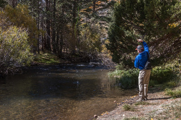 Obraz premium Fly fisherman fishing in a small beautiful stream in the mountains. He's hooked a small fish. He is wearing a blue jacket and a ball cap. He has a small pack on his back. His line is bright green. 