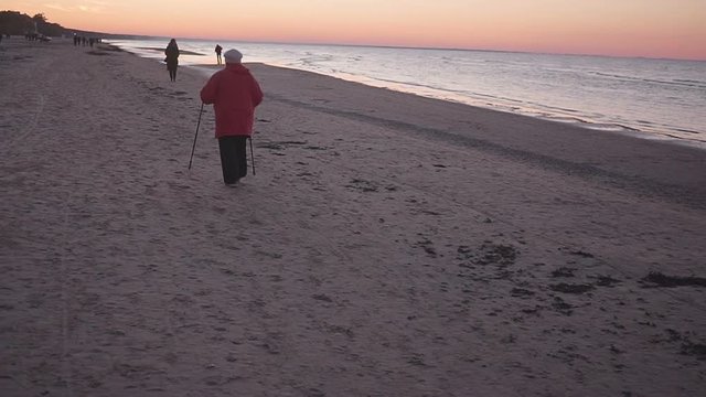 Old woman goes with Nordic walking sticks along the seashore.