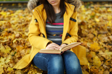 Obraz premium Woman in yellow coat and jeans sitting under the maple tree with a red book in fall city park on a warm day. Autumn golden leaves. Reading concept. Close up.
