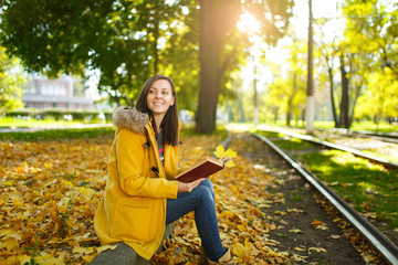 A beautiful happy brown-haired woman in a yellow coat and jeans sits alone in the park near the tram tracks and reads a book in the fall warm day. Autumn yellow leaves.