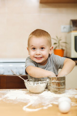 Little kid boy helps mother to cook Christmas ginger biscuit in light kitchen. Happy fair-haired child in gray T-shirt 2-3 years at the table with iron sieve and flour in weekend morning at home.