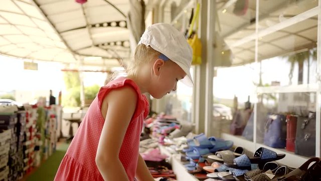 little girl is considering and choosing summer flip-flops in a small street market in sunny day