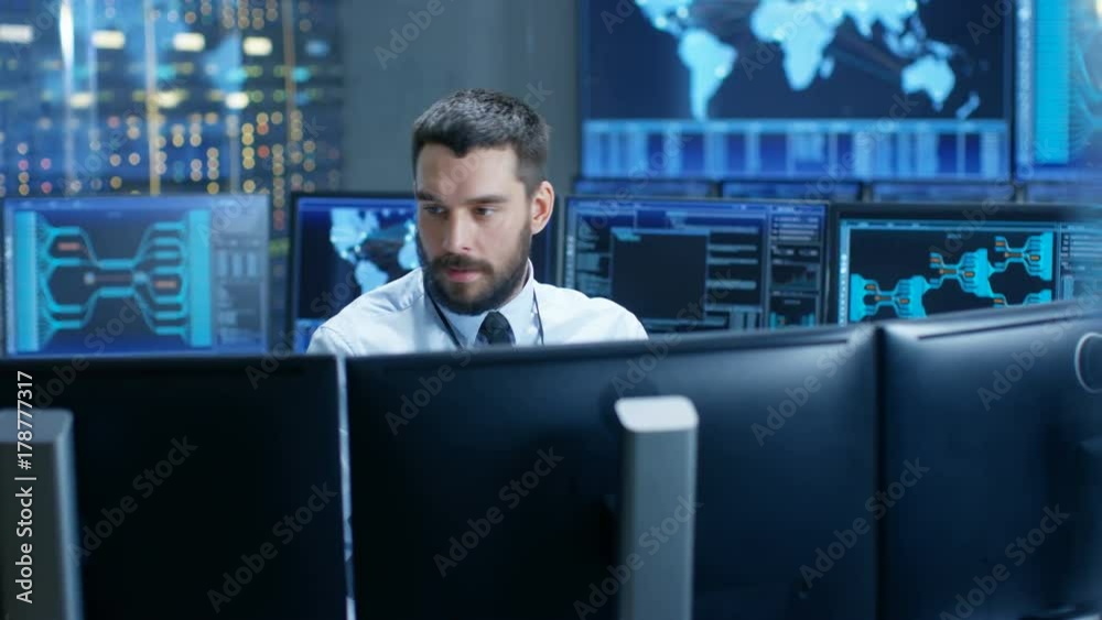 Fototapete In the System Control Center Technical Operator Monitors Displays at His Workstation. He's Surrounded by Displays Showing Relevant Data. In the Background Data with Interactive Map. 