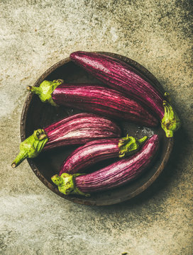 Flat-lay of fresh raw Fall harvest purple eggplants or aubergines in wooden bowl over concrete stone background, top view. Healthy Autumn vegan cooking ingredient