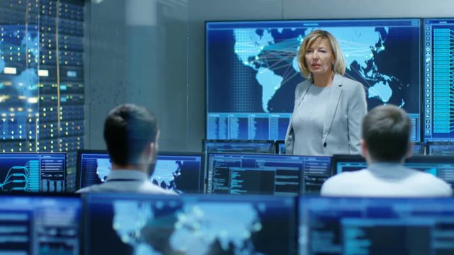 In the System Monitoring Room Senior Female Engineer Holds Briefing For Her Subordinate Specialists, Explains Their Goals and Tasks. They Work in the High-Tech Environment. 