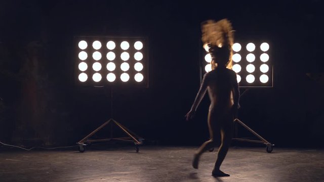 a young woman of pleasant appearance dances in the training hall in the lighting of searchlights in the background. the girl professional dance performs the set number in the style of classical and