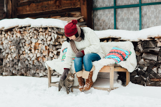 Woman And A Cat Relaxing Outside A Country House On A Cold Winter Day. Portrait Of A Woman Stroking A Cat In The Snow. 