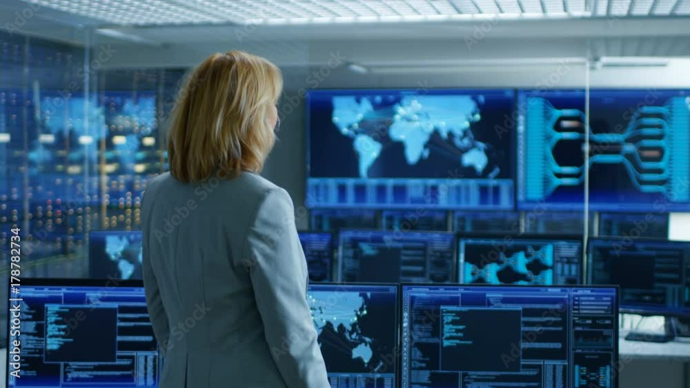 Fototapete In the System Control Room Female Head of the Department Crosses Arms and Thinks Standing with Her Back to the Camera. In the Background Multiple Screens Showing Interactive Data. 