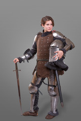 young woman in Knight Armour holding helmet in one hand and sword on her shoulder over grey...