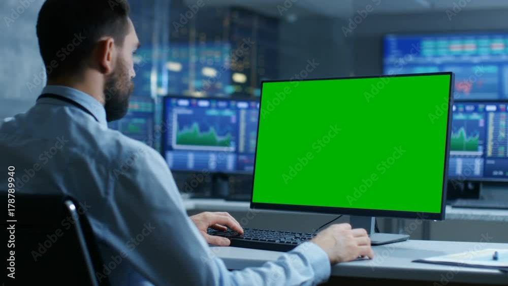 Poster Over the Shoulder View of Stock Market Trader Working on a Computer with Isolated Mock-up Green Screen. In the Background Monitors Show Stock Ticker Numbers and Graphs. 