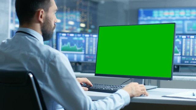 Over the Shoulder View of Stock Market Trader Working on a Computer with Isolated Mock-up Green Screen. In the Background Monitors Show Stock Ticker Numbers and Graphs.