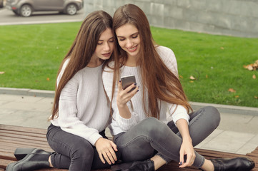 Two girlfriends are sitting on the bench and watching pictures in the smartphone