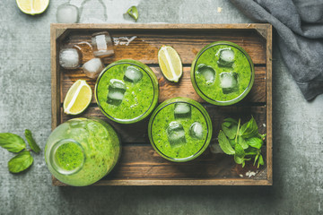 Fresh green smoothie in bottle and glasses with ice cubes, mint and lime in wooden tray over grey concrete background, top view. Clean eating, detox, vegetarian, weight loss, raw, healthy food concept