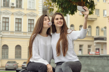 Two girlfriends puff bubbles out of chewing gum while they make a selfie photo