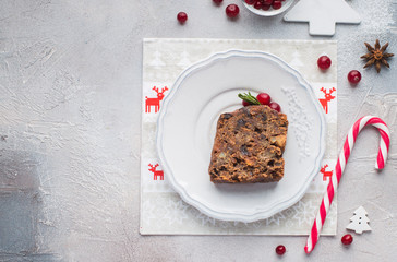 A piece of traditional Christmas English pudding (fruit cake) on white plate on a concrete light background. Christmas decor of the table. Funny napkins with reindeers Santa Claus.  Flat lay, top view