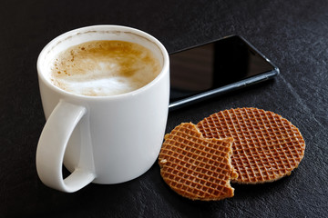 Milky frothy coffee in white mug partially drunk next to partially eaten waffle  biscuits isolated on black.