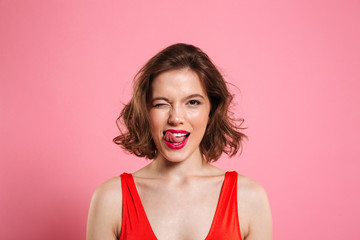 Close-up portrait of pretty playful woman winks one eye, showing tongue, looking at camera, isolated on pink background